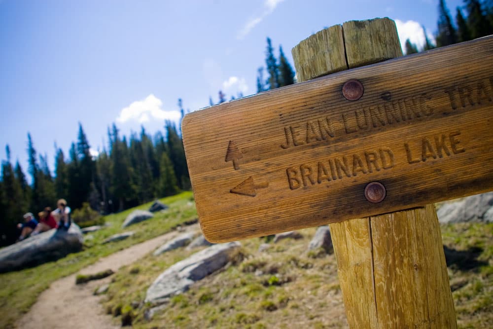 A tilted angle of the wooden trail marker has two arrows with the names of trails on it. In the background people sit on a bench surrounded by evergreen trees and grasses.