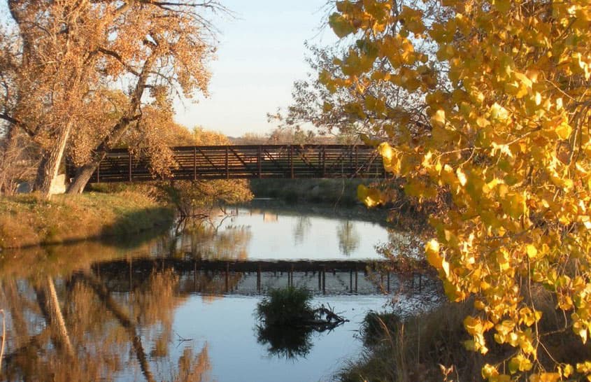 A pedestrian bridge sits over a calm river with golden leafed trees to the right and left.