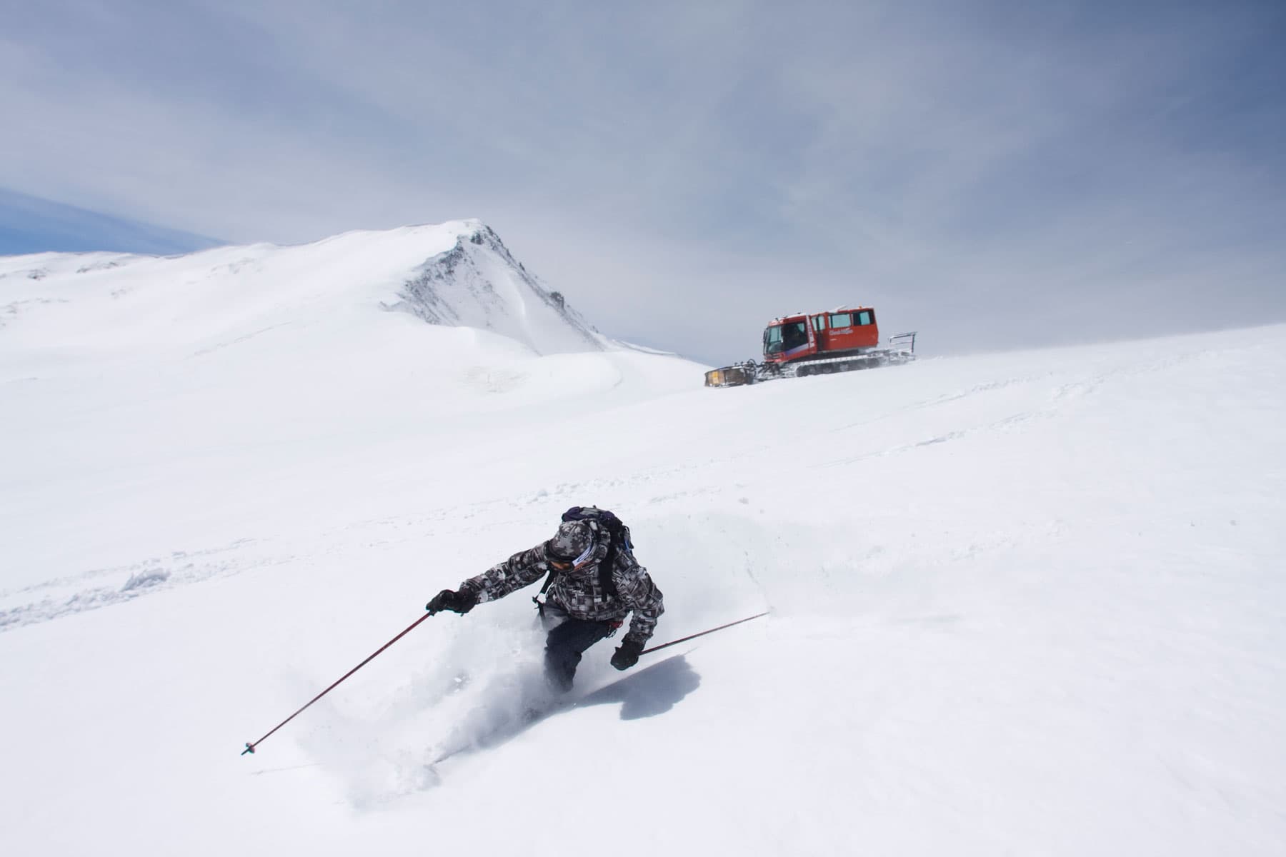 A skier makes their way down a snowy slope with a red snowcat sitting atop the ridge in the distance.