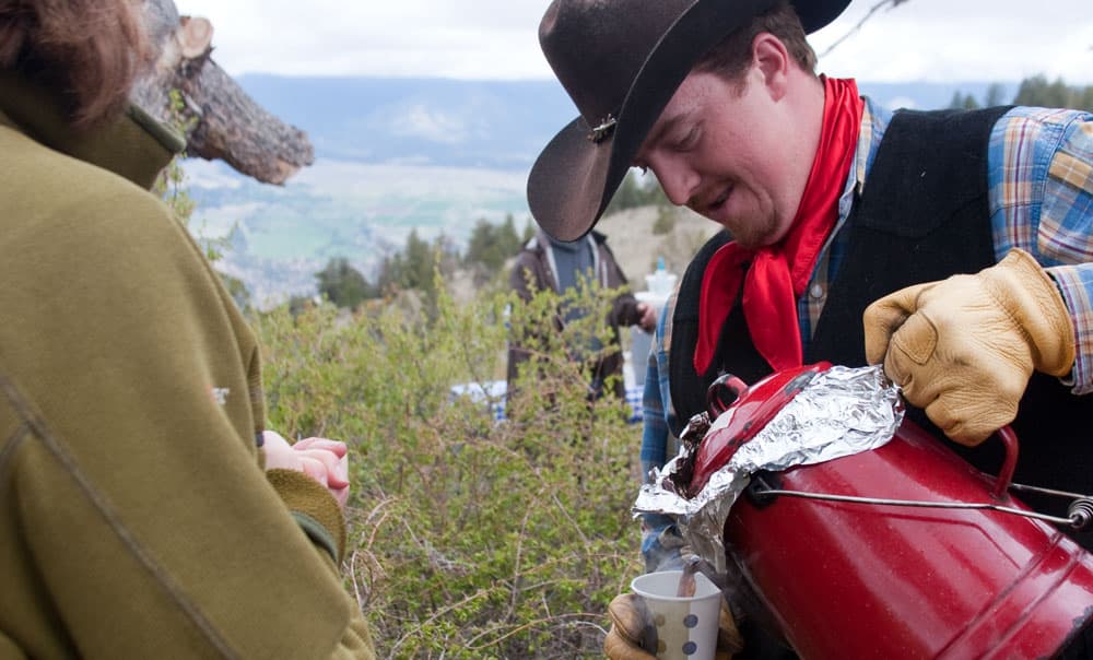 A cowpoke in a brown leather hat, black vest and shiny, red ascot pours steaming hot coffee from a red coffee pot for a guest at a Colorado dude ranch.