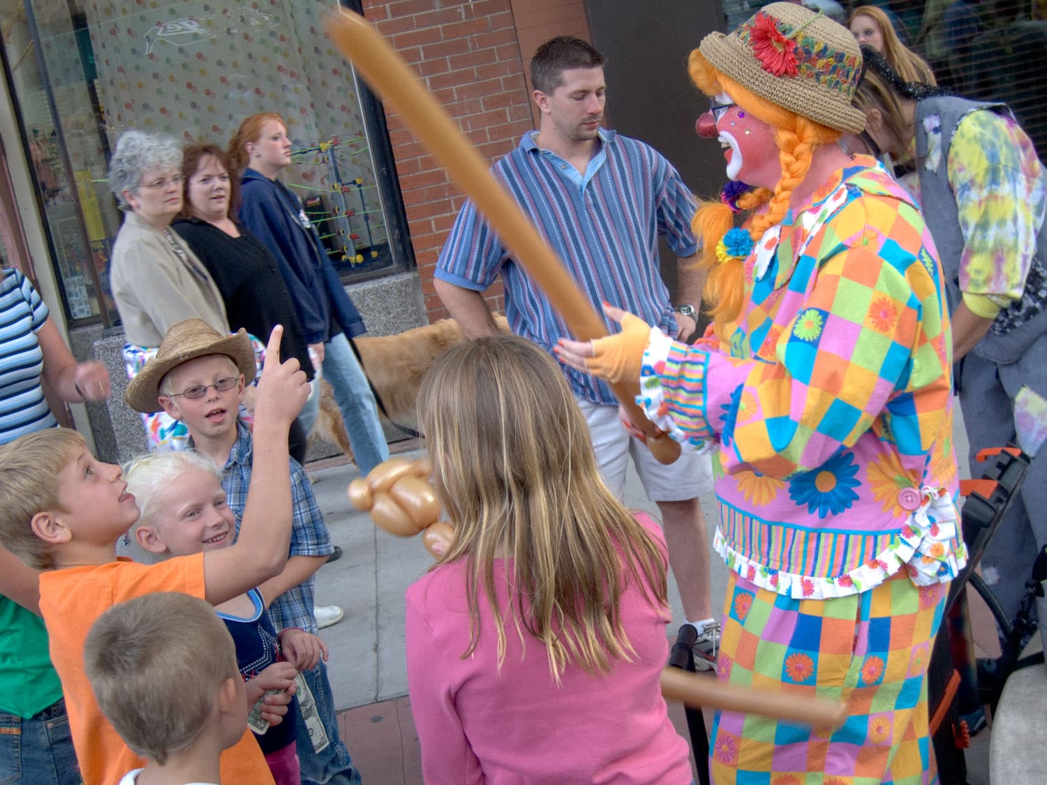 A clown with orange, braided pigtails and a colorful, checked outfit makes balloon animals for a group of children at the Grand Junction Farmers' Market.