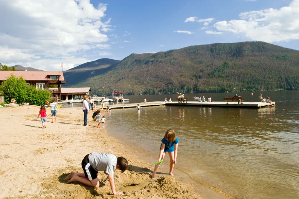 Several people sit on benches or dangle their legs off the edge of a floating, wooden dock on Grand Lake in Colorado. On the shore, children play in the sand or wade in the shallow waters.