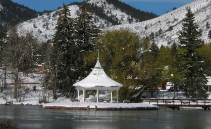 The white historic gazebo in Green Mountain Falls sits on a lake. The ground is covered in snow and the evergreen trees are dusted with snow. Behind them there are snow-covered mountains.