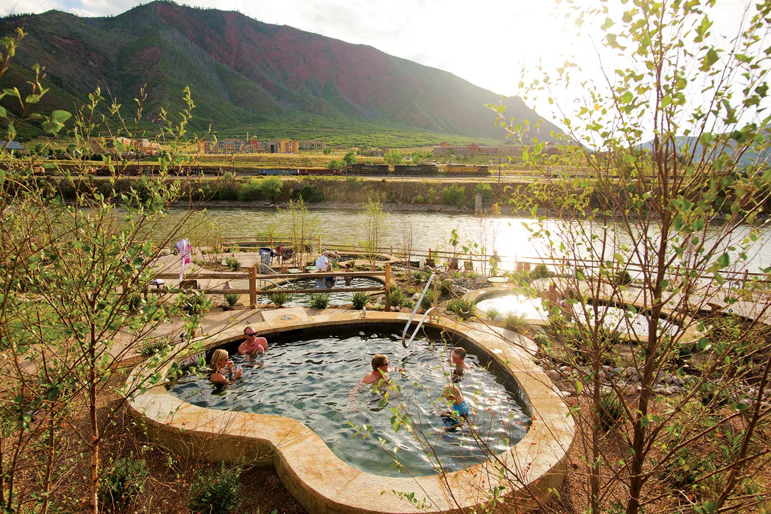 A group of people soaks in a stone hot pool overlooking a river and the mountains of Iron Mountain Hot Springs in Glenwood Springs. The sun glimmers off of the water.