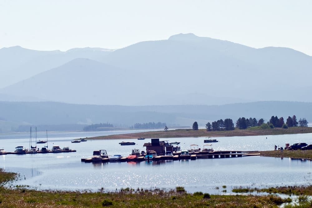 Boats are docked on a sparkling Lake Granby with hazy Rocky Mountains in the background.