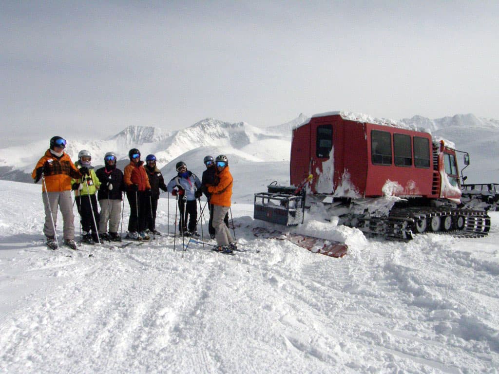 A group of skiers looks at the camera next to a red snowcat on a snowy landscape. Snow-covered mountains dot the background and the sky is white.