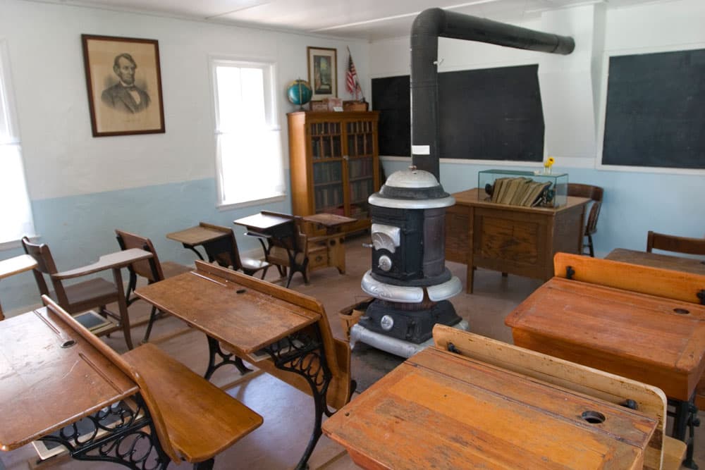 A large, antique stove rests in the middle of an old school room at a museum in Colorado. The room is filled with antique desks and other artifacts.