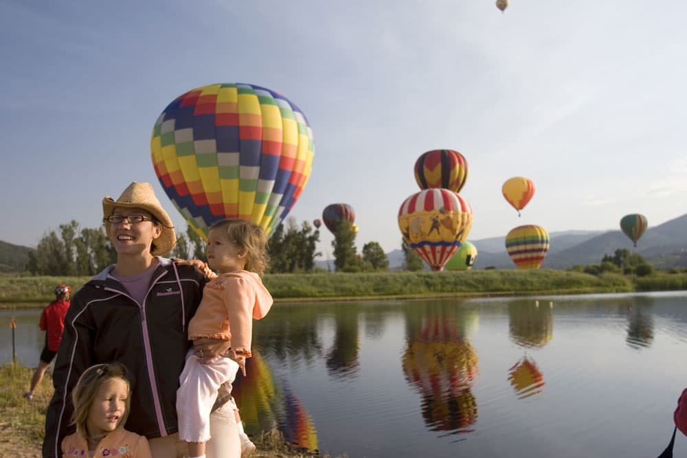 A parent stands with two young children on the banks of a tranquil body of water in Steamboat Springs, Colorado. Behind them, massive hot air balloons of different colors and designs rise into the air.