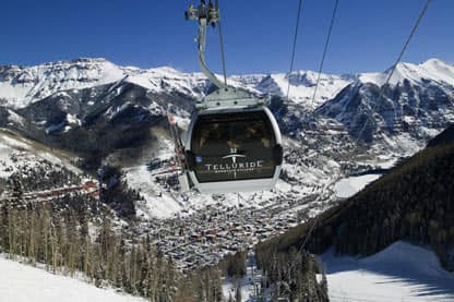 A gondola with Telluride's logo climbs its way up the cables away from the downtown area. Above town, blue mountain peaks are covered in snow.