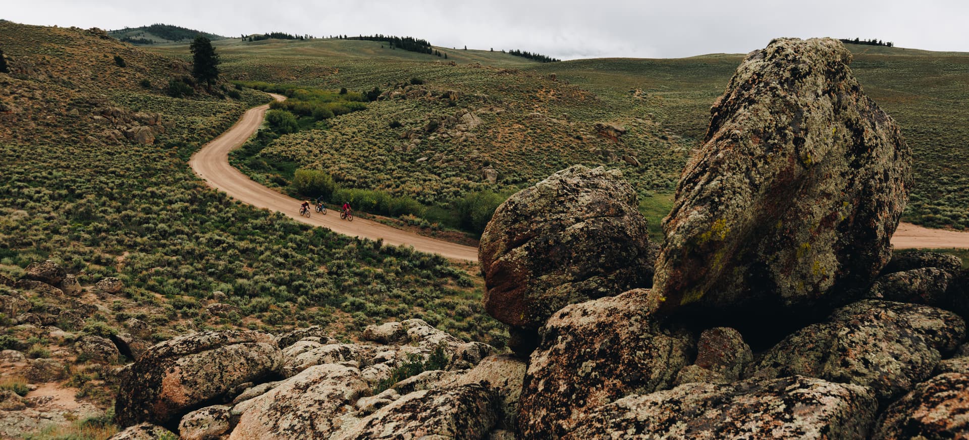 People on mountain bikes ride a trail through green hills and rocky terrain in Gunnison Crested Butte in Colorado