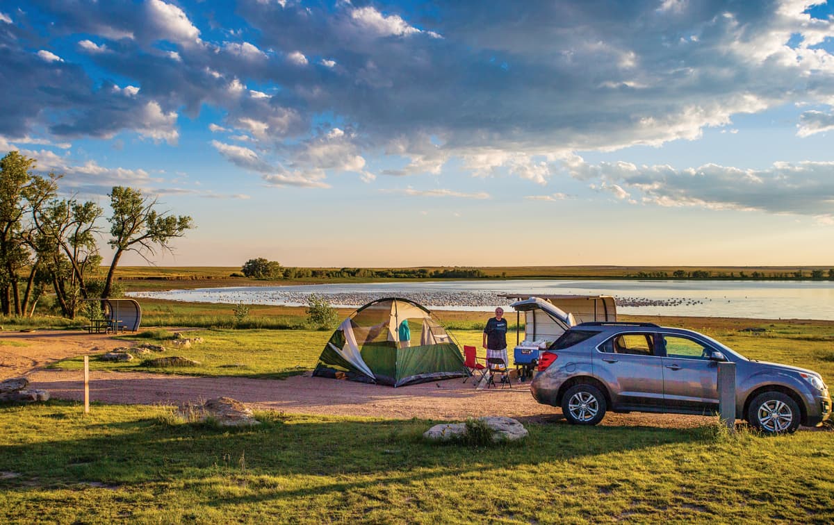 A tent and a car in front of a lake and trees while camping at North Sterling State Park in Colorado