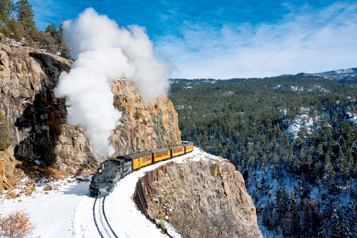 A black train engine billows steam as it travels down the Durango & Silverton Narrow Gauge Railroad in winter. It pulls several yellow passenger cars around a cliff.
