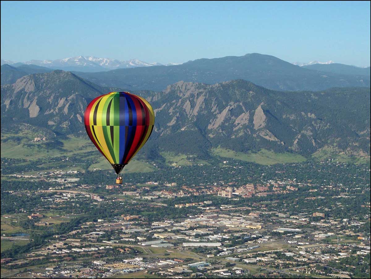 A red, blue, green, yellow and black hot air balloon floats high over a town at the base of a forested mountain in Colorado.