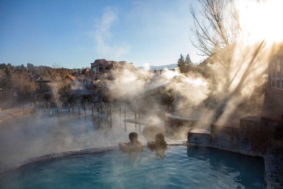 Two people are chest-deep in a warm pool overlooking more pools under a blue sky. The steaming pool is part of the Springs Resort & Spa in Pagosa Springs.