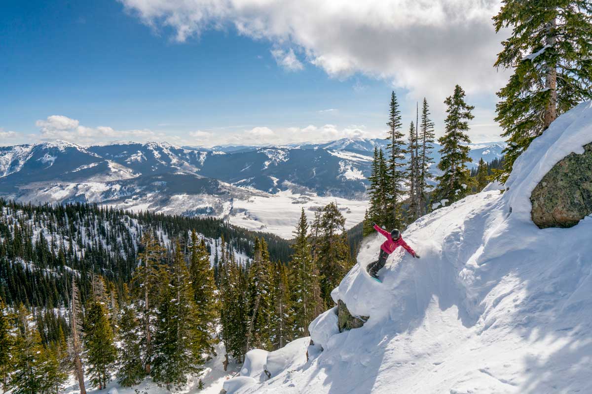 A downhill skier carves a turn over a rock face on what looks to be expert backcountry terrain; the sun is shining and we can see an infinity of mountain peaks covered in snow