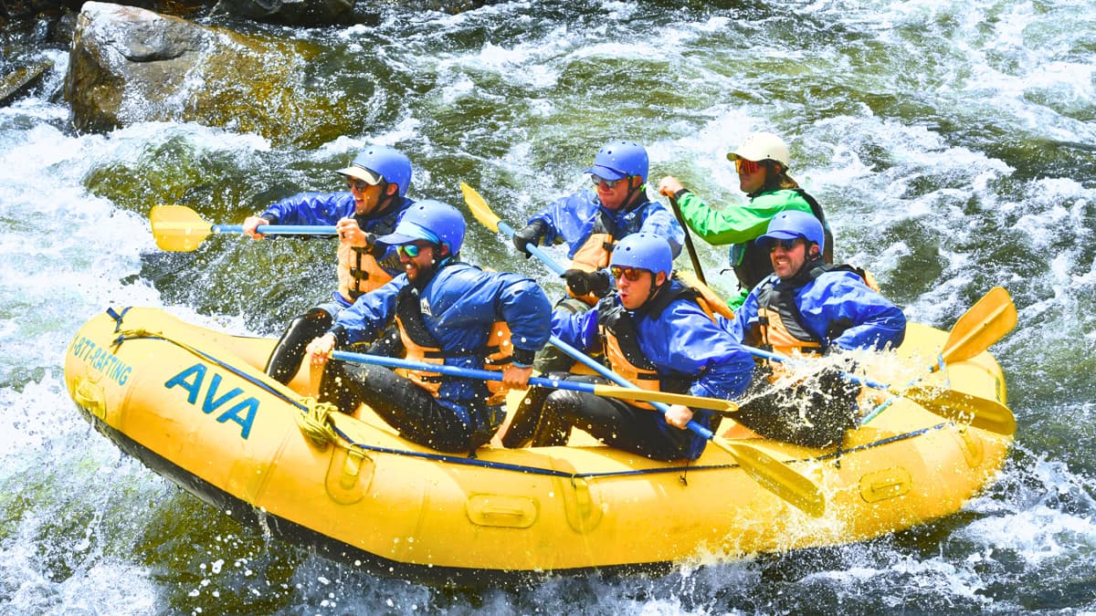 Six people smile and laugh on an AVA Rafting & Zipline whitewater rafting adventure. They are all in blue helmets except for the guide in the very back, who looks calm and collected. They all have paddles in their hands as they ride the waves.