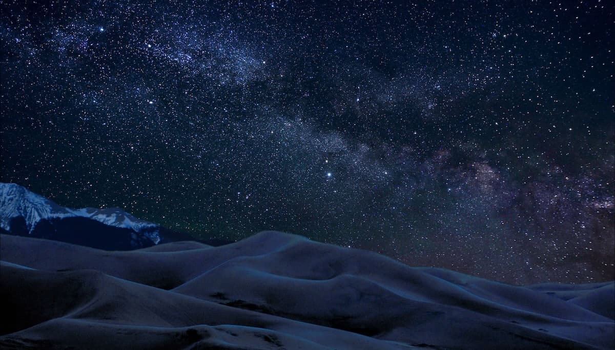 A dark blue and purple sky with the Milky Way over large sand dunes
