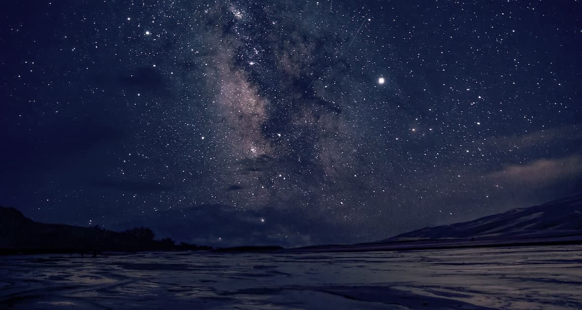 Hundreds of stars in a dark-blue night sky over mountains and sand