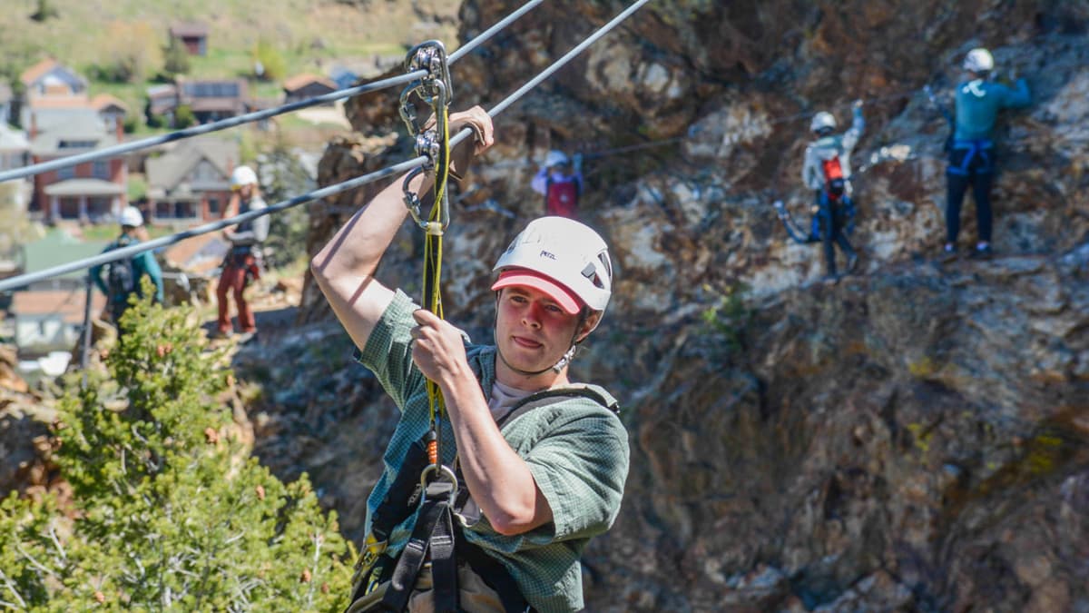 Someone in a hat and helmet slides down the zipline at AVA Rafting & Zipline in Buena Vista. There are four people behind this person climbing in the background. The sun is shining.