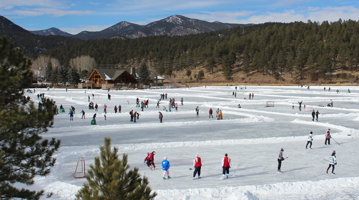 People in hockey and skating gear enjoy themselves on the white rink of the Evergreen Lake in the winter. In the background are mountains and groves of pine trees, underneath mostly blue skies.