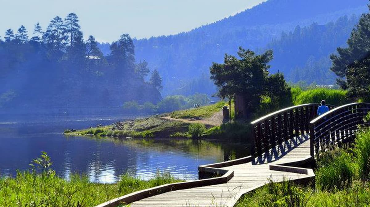 A winding wooden path goes over the blue waters of the Evergreen Lake, surrounded by mountains and groves of green pine trees.