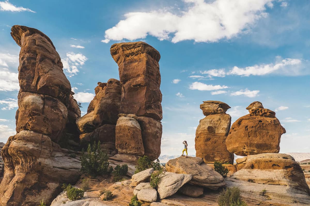 Someone in athletic wear stands looking out at the scene at Devil's Kitchen in Colorado National Monument in Grand Junction, Colorado. Around the person stand towering stone formations that make up the Devil's Kitchen. The sky above is peppered with wispy white clouds, but mostly blue.