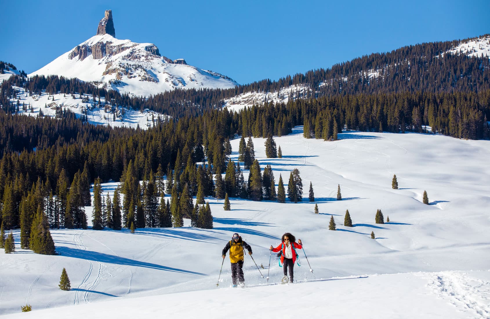 Two people cross a snowy field under blue skies on snowshoes