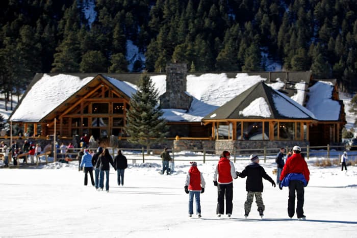 In the foreground, a family of four on ice skats holds hands as they glide toward a wooden lodge with snow on the roof. Other skaters can be seen in the background.