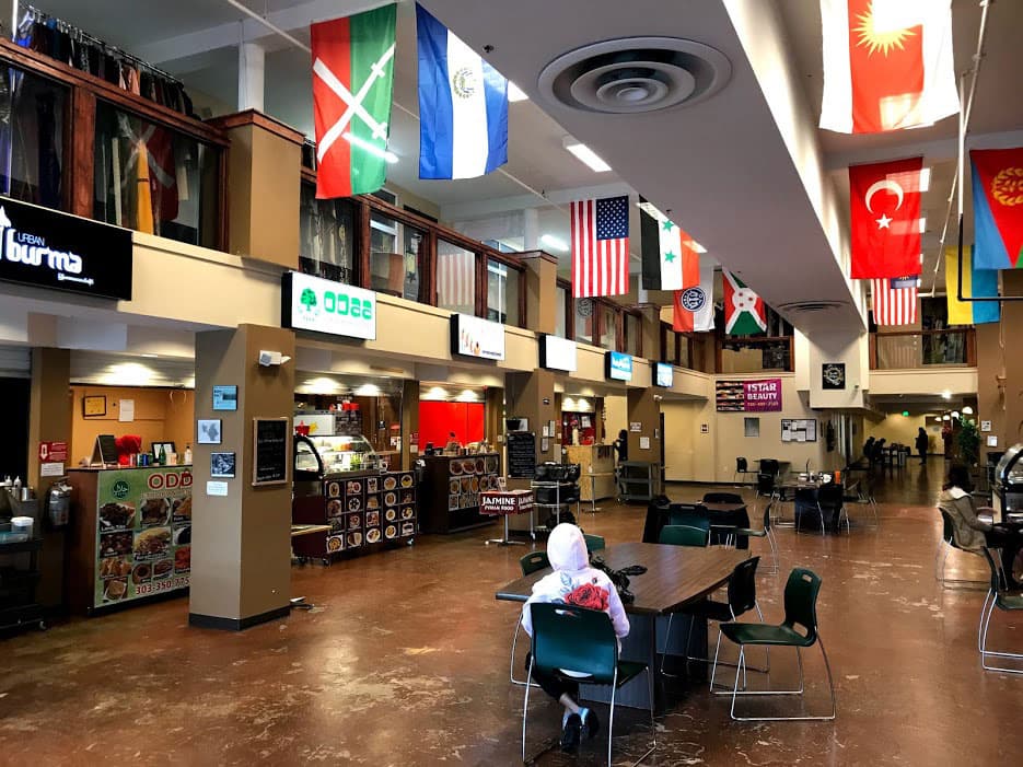 A lone diner sits at a table waiting for food at the food court at Mango House in Colorado. Above them, numerous international flags represent some of  the diverse cuisines present.