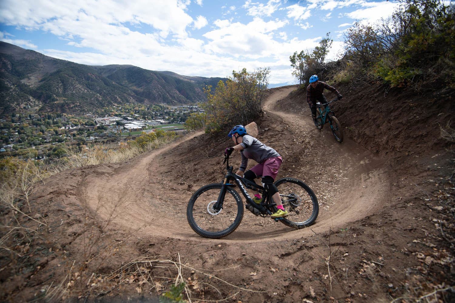 Two mountain bikers wearing helmets and knee pads whip around a tight, switchback curve on a mountain trail near Glenwood Springs, Colorado.
