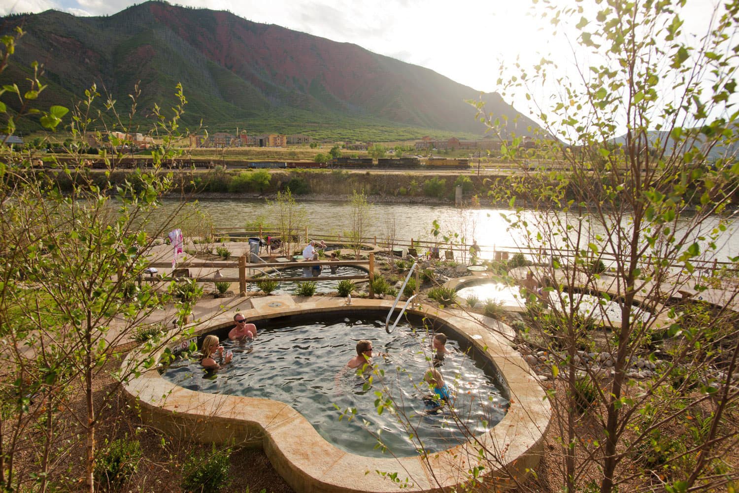 Families and couples soak in sandstone-covered pools next to a large body of water and amid saplings with new spring leaves at Iron Mountain Hot Springs in Colorado.