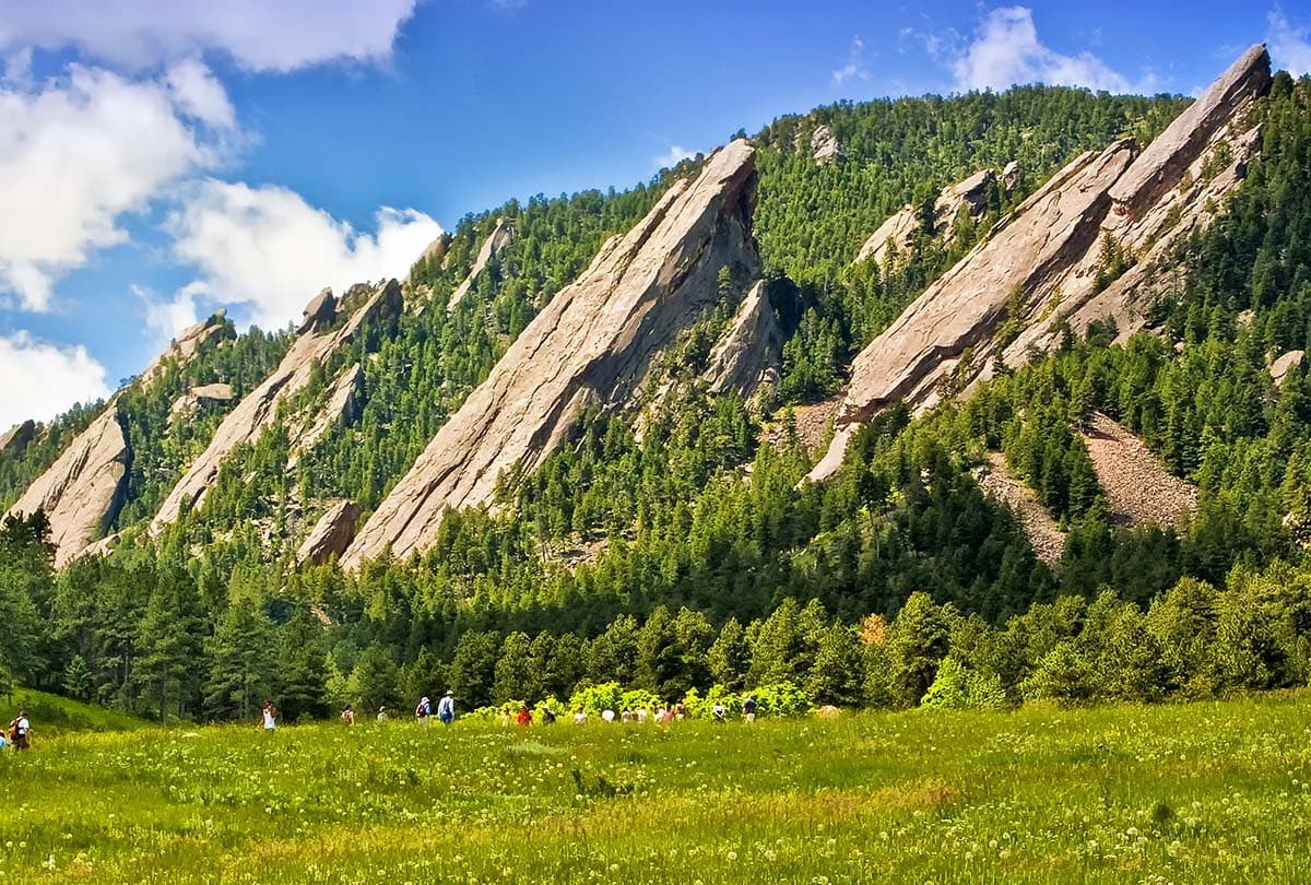 Several people walk the grassy foothills near Boulder, Colorado, and gather before a steep, imposing mountainside, featuring a thick forest and raised, angular slabs of gray rocks.