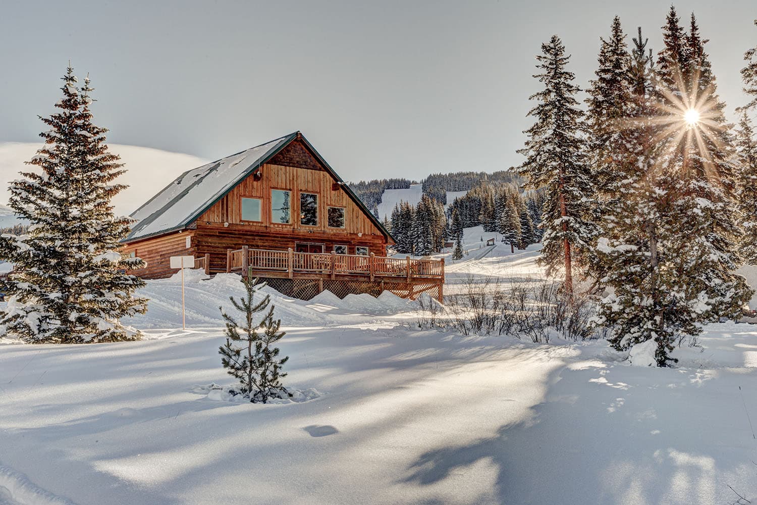 The sun glimmers through evergreens next to a wood ski lodge surrounded by blankets of snow