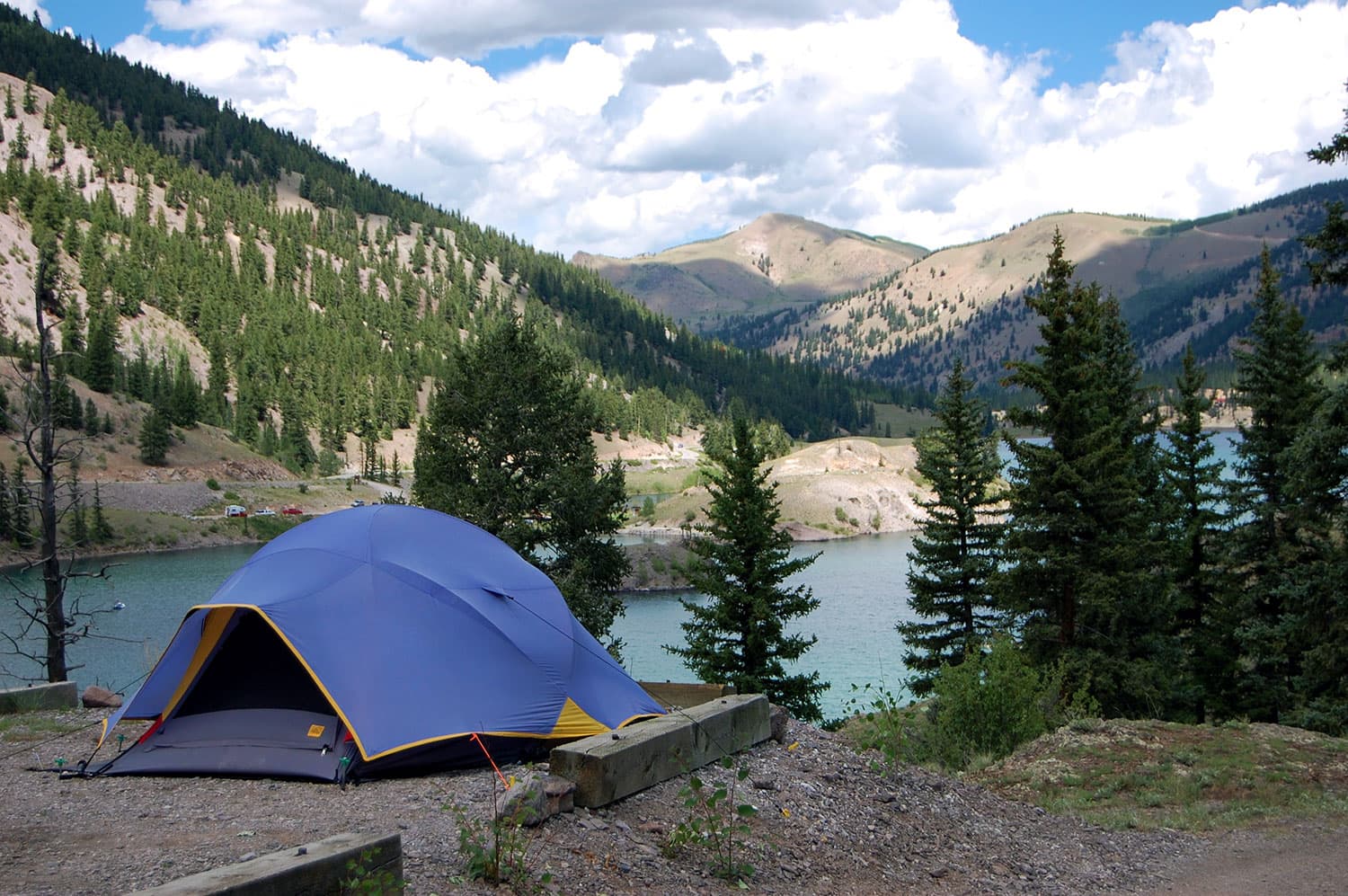 A blue tent is pitched on a campsite next to a lake surrounded by evergreen trees