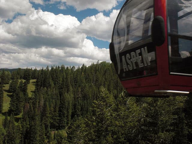 Aspen Mountain's red gondola flies high above a field of evergreens. Its glass, which has printed on it "ASPEN," reflects clouds in a blue sky.