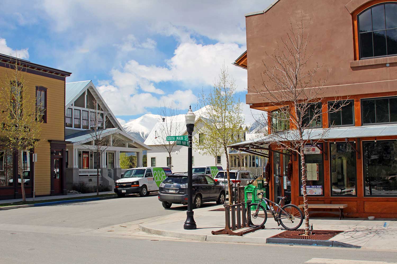 A bike sits in a bike rack on a corner sidewalk outside a store lined with windows in downtown Buena Vista, Colorado.  Houses and stores line the other side of the street.