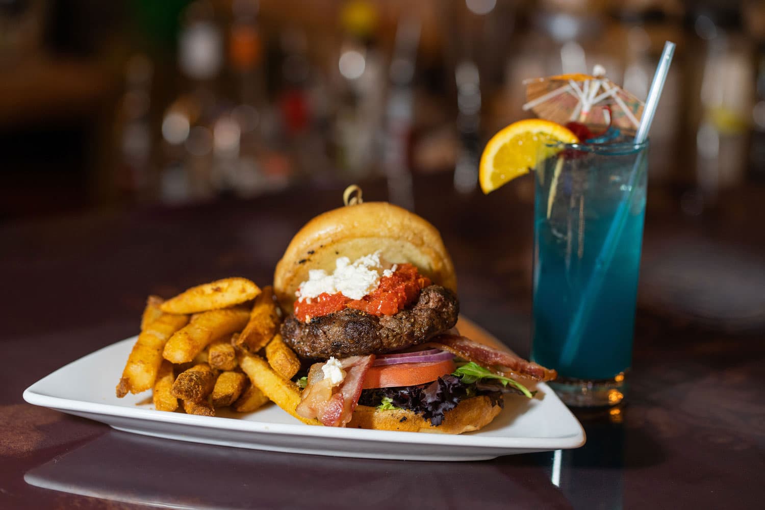 A juicy burger and blue cocktail with a lemon wedge sit on a table