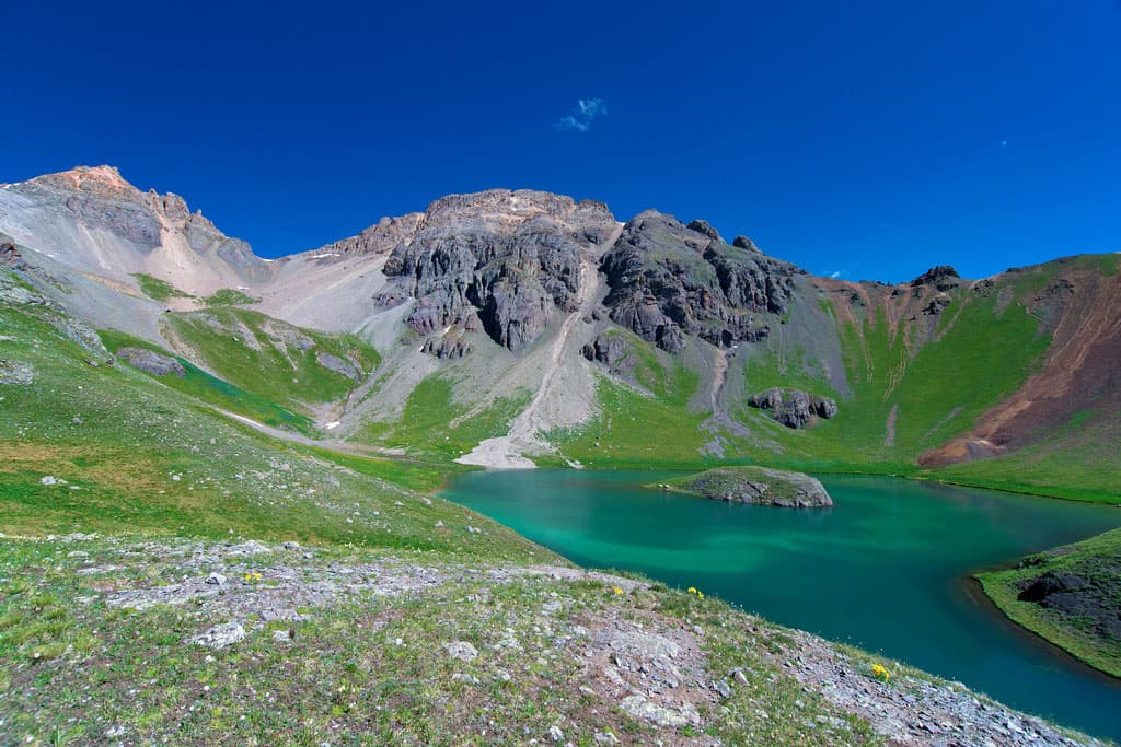 Turquoise waters of a lake surround a small rock island near Silverton, Colorado. The lake lies at the base of steep grassy hills on multiple sides.