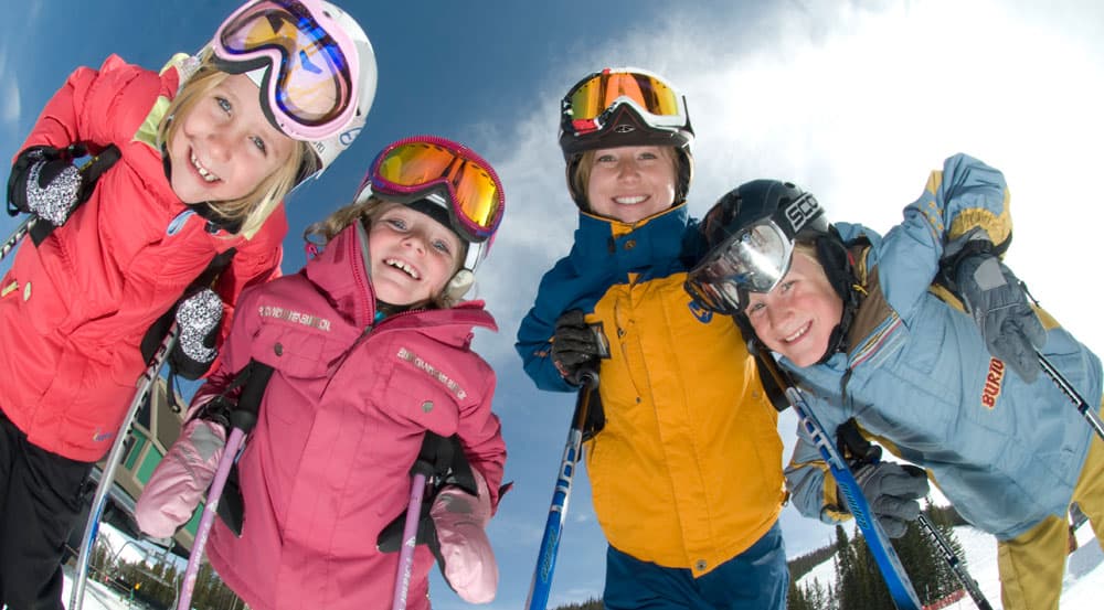 Two adults and two kids in ski gear stand over the camera with the sun behind them