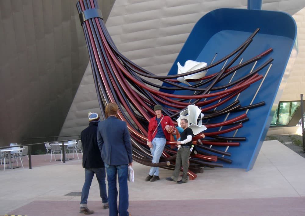 People pose in front of a giant sculpture of a blue dust pan and blue and red whisk sweeping crumpled paper at the Denver Art Museum.