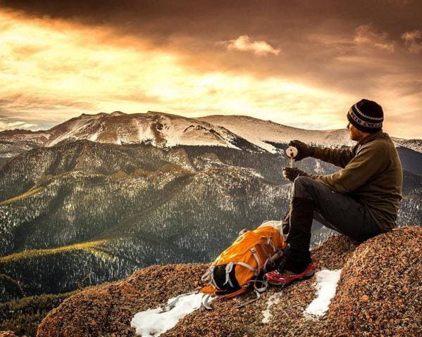 A hiker, bundled in warm clothes and a hat, sits next to his orange backpack on the side of a hiking trail. In the distance, we gazes a snow-covered mountains.