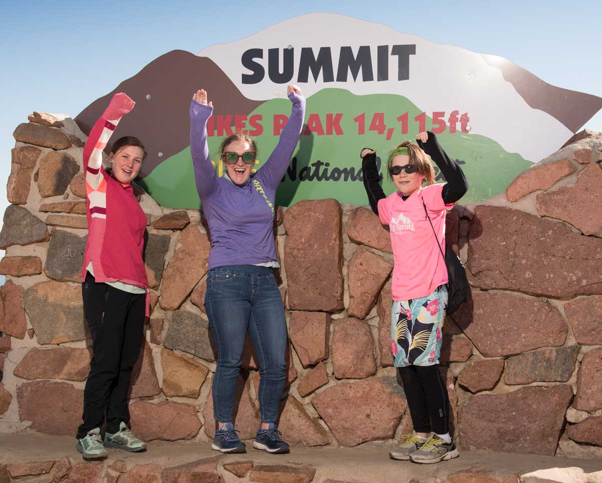 Three riders celebrate with their hands in the air in front of a sign at Pikes Peak's summit