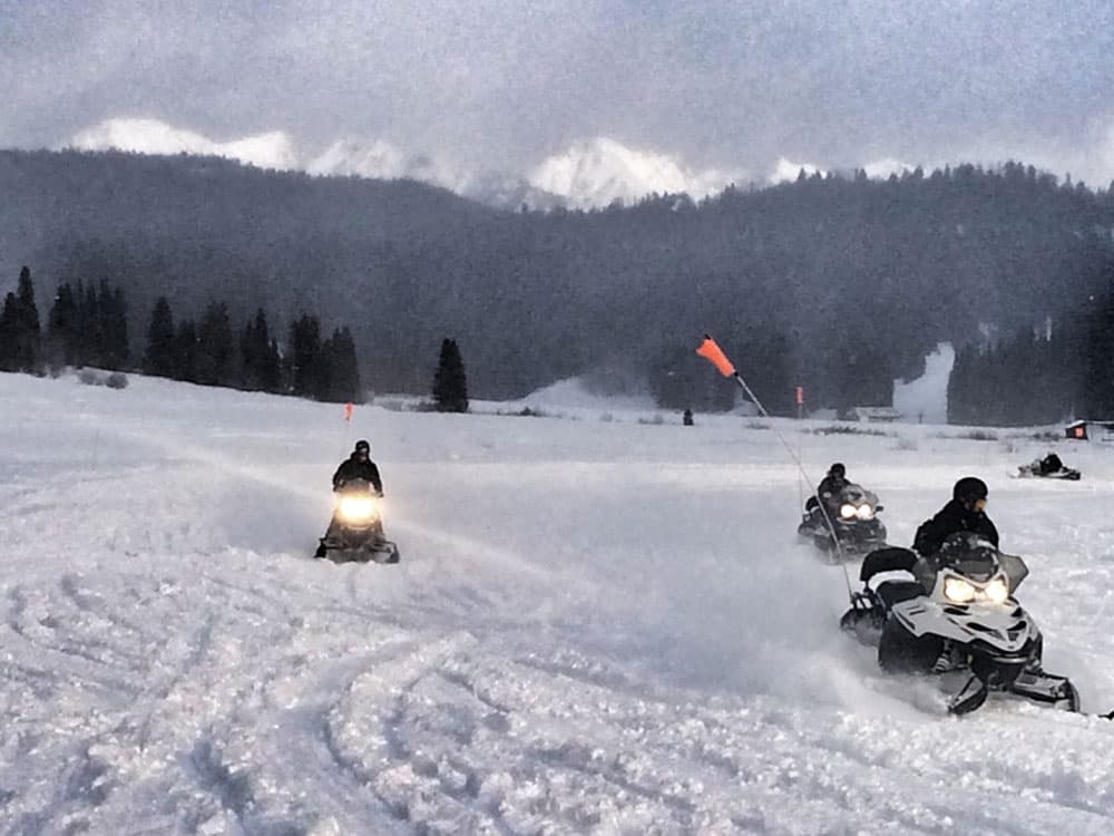 Snowmobiles glide across the snow with trees and mountains in the background