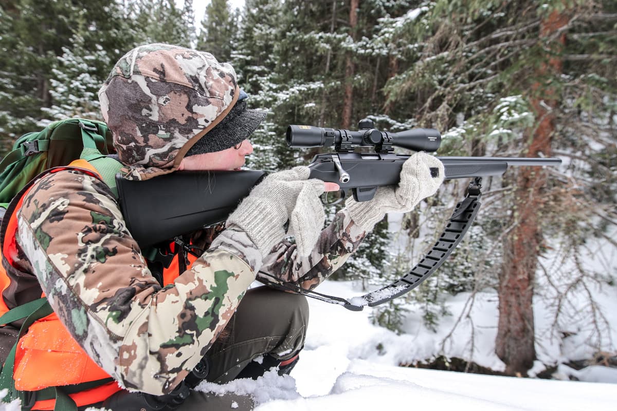 A hunter in camouflage and an orange vest peers through a rifle scope