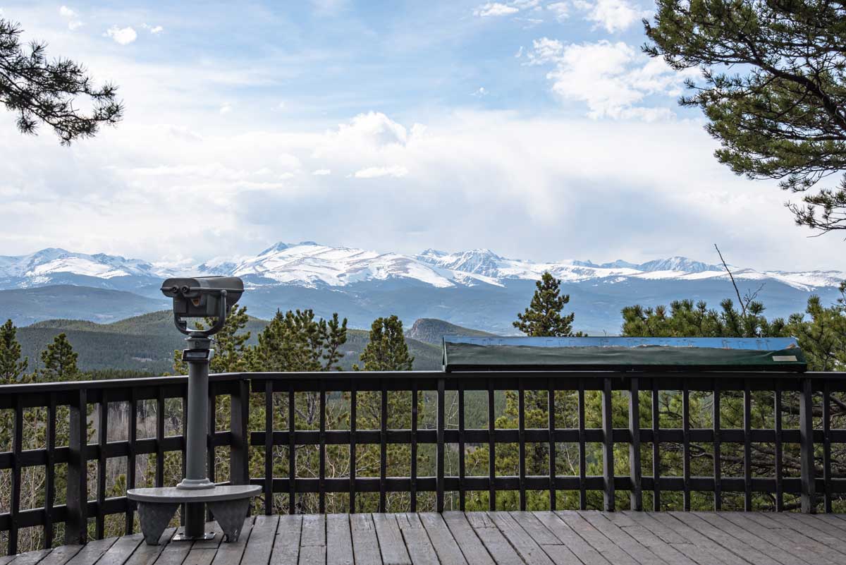 A wooden viewing platform offers panoramic views of the forest and snow-capped mountains of Golden Gate Canyon State Park.