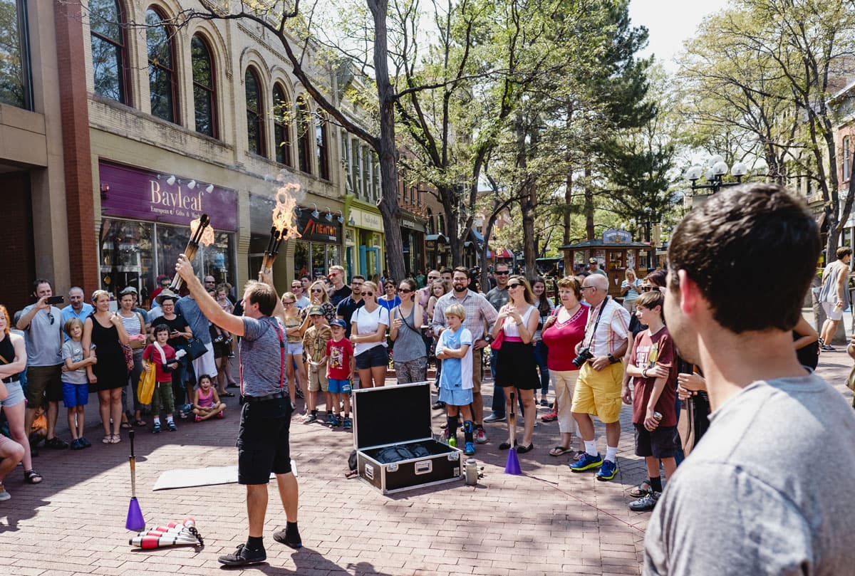 A crowd gathers around a street performer on Pearl Street in Boulder. The performer displays several flaming batons over their head.