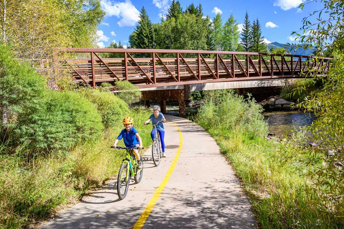 Kids biking on a paved path near a river with a bridge in the background. They're surrounded by green trees and shrubs.