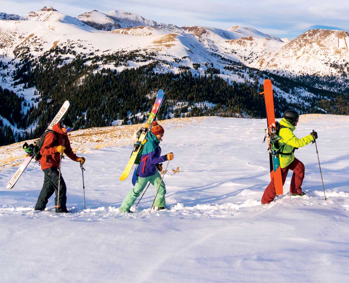 Three people wearing colorful snow gear and hauling skis walk through glistening snow on Loveland Pass. Behind them is a towering mountain covered with snow and pine trees.