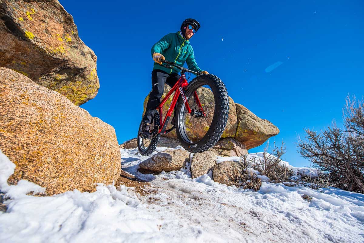 A fat biker and his meaty tires jumps between two rocks onto a snoy trail. The sky is bright blue above him