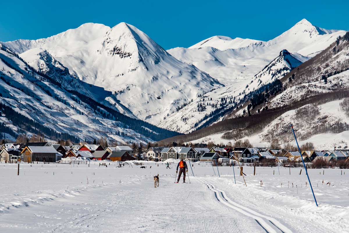 Person Nordic skiing on a groomed trail in Crested Butte with a dog trotting beside them. There are bright blue skies contrasted with a series of snowcovered peaks.